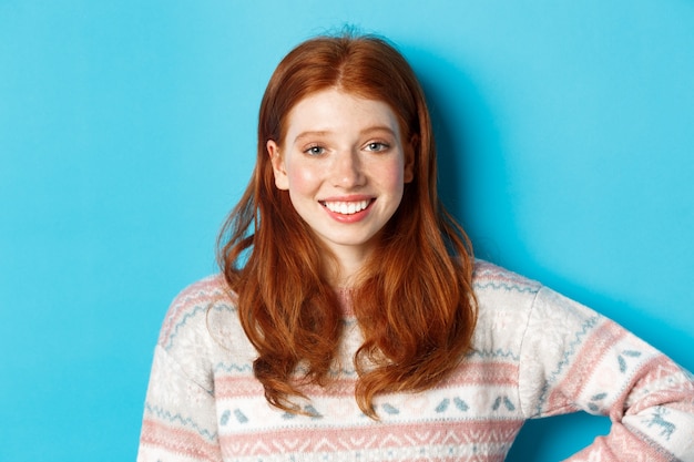 Close-up of attractive female student with red hair, wearing winter sweater, smiling happy and staring at camera, blue background.