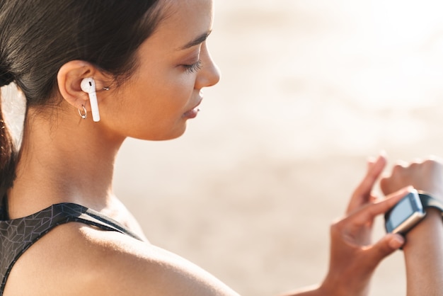 Close up of an attractive fit sportswoman outdoors, looking at smartwatch while sitting