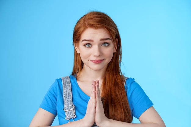 Close-up attractive tender redhead woman asking pretty please, supplicating, hold hands pray, namaste gesture, look sincere pity, smiling begging help favor, stand blue background