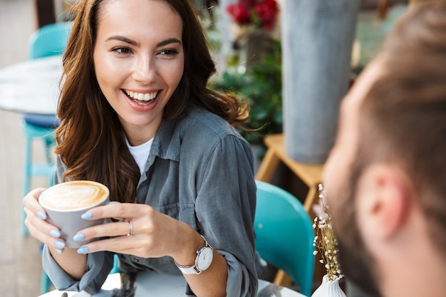 屋外のカフェのテーブルに座って、コーヒーを飲みながら、おしゃべりしながら昼食をとるのが大好きな魅力的な若いカップルのクローズアップ