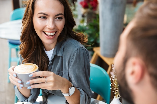 Photo close up of an attractive young couple in love having lunch while sitting at the cafe table outdoors, drinking coffee, talking