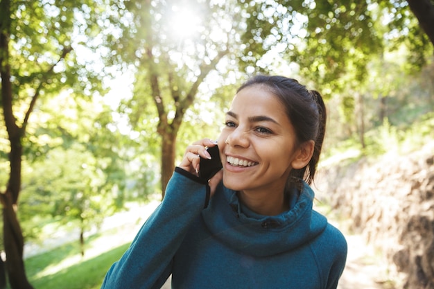 Close up of an attractive young fitness woman wearing sportswear exercising outdoors, using mobile phone