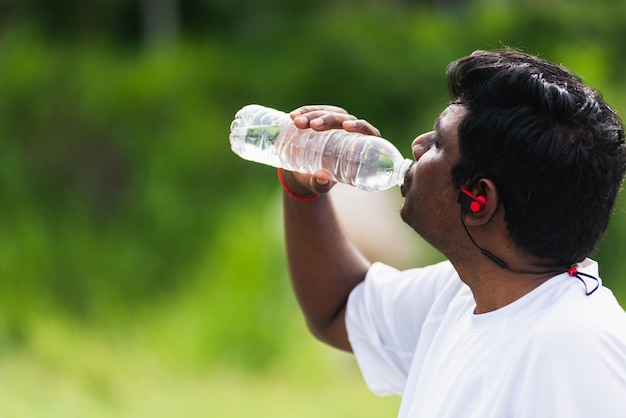 Close-up Aziatische jonge sport runner zwarte man draagt atleet koptelefoon hij drinkt water uit een fles na het hardlopen in het outdoor street health park, gezonde oefening training concept