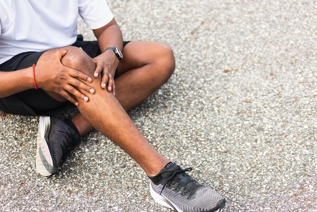 Close-up Aziatische sportloper zwarte man draagt horloge zittend hij gebruikt handen gezamenlijke greep op zijn knie terwijl hij rent in het buitenstraatgezondheidspark, gezonde oefening Verwonding door trainingsconcept