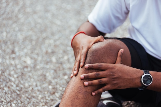 Foto close-up aziatische sportloper zwarte man draagt horloge zittend hij gebruikt handen gezamenlijke greep op zijn knie terwijl hij rent in het buitenstraatgezondheidspark, gezonde oefening verwonding door trainingsconcept