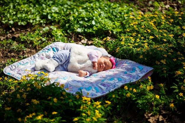 Крупным планом ребенок с бантом на голове лежит на белом пледе на зеленой траве. На улице весна, на ребенка светит солнышко