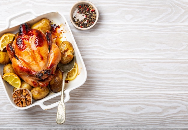 Primo piano di pollo biologico croccante intero al forno o arrosto con patate, aglio, limone in casseruola bianca con cucchiaio da portata su sfondo bianco in legno, vista dall'alto e spazio per il testo