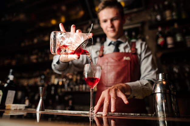 Chiuda sul barista che versa il cocktail rosso luminoso dell'alcool in vetro operato