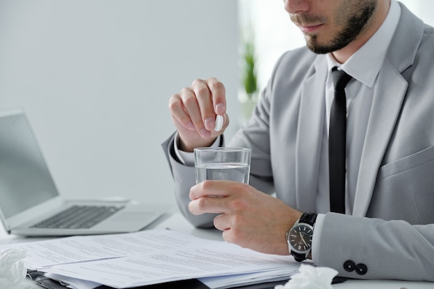 Close-up of bearded businessman sitting at desk with papers and laptop and taking hangover prevention pill in office