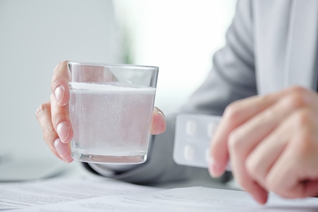 Close-up of bearded businessman sitting at desk with papers and laptop and taking hangover prevention pill in office