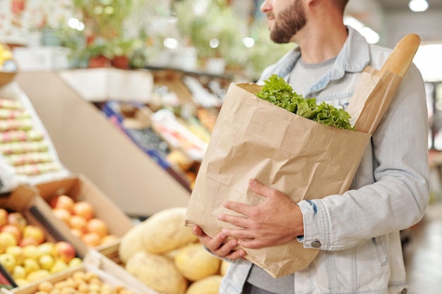 食品市場で新鮮な製品の完全な紙袋を運ぶデニムジャケットのひげを生やした男のクローズアップ