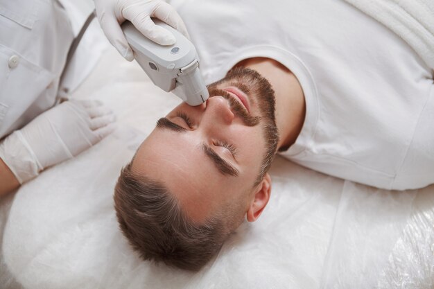 Close up of a bearded man enjoying getting laser facial treatment by professional cosmetologist