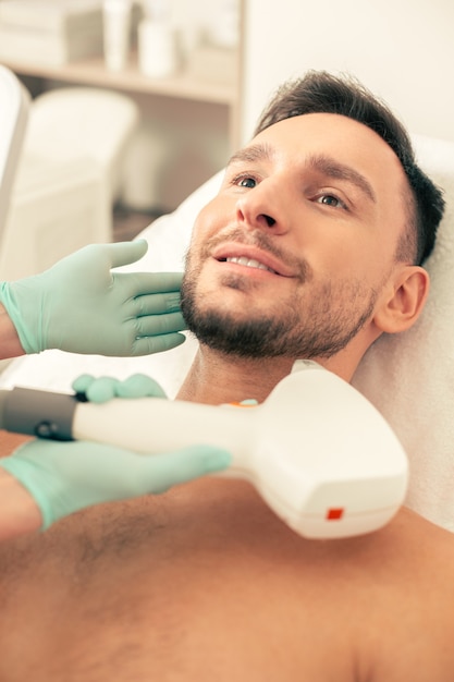 Close up of the bearded smiling man looking glad while having modern tool near his face during the laser skin procedure