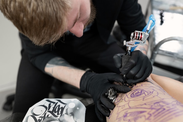 Close up of the bearded tattoo male artist makes a tattoo on a female leg