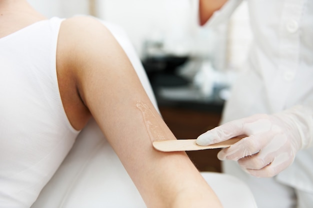 Close-up. The beautician holds a wooden spatula and applies a transparent gel with anesthetic to the woman's hand and prepares the skin for the laser hair removal procedure.