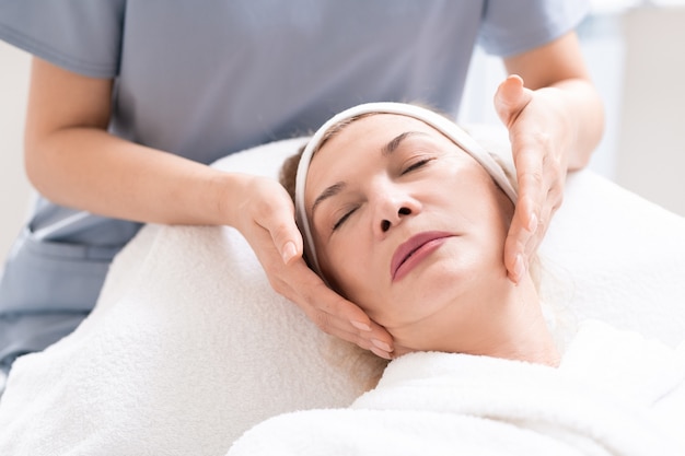 Primo piano dell'estetista che massaggia il fronte del cliente femminile maturo che si rilassa alla procedura