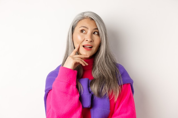 Photo close up of beautiful asian elderly woman in stylish clothes, making plans, looking aside and thinking, standing on white.