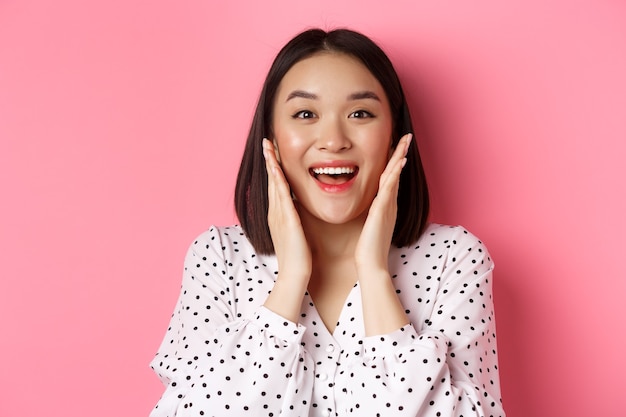 Close-up of beautiful asian woman touching clean skin face and staring at camera amazed, standing over pink