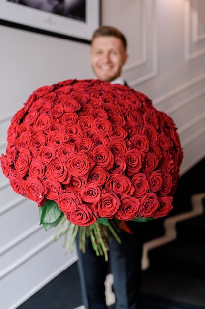 Close up of beautiful big bouquet of flowers red roses which holding smiling fiance man standing on stairs indoor Gift for a loved one Valentine's day