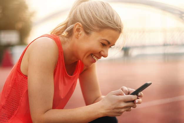 Photo close up of beautiful blonde woman with ponytail and dressed in sportswear typing message on smart phone. fitness outdoor concept.