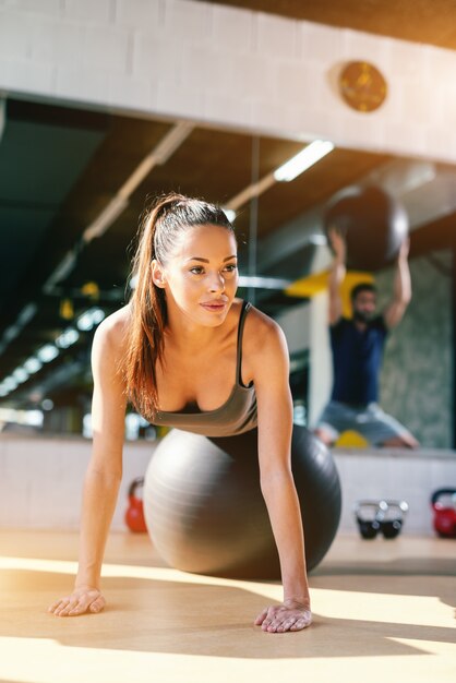 Chiuda in su di bella donna caucasica con coda di cavallo e lunghi capelli castani facendo esercizi con palla di pilates in palestra. in background riflesso dell'uomo con palla pilates.