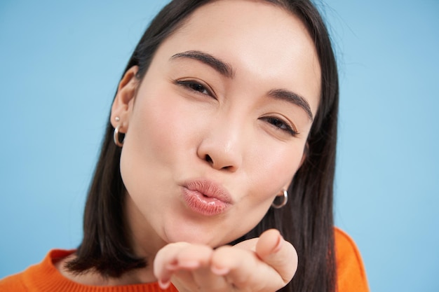 Close up of beautiful korean woman blowing air kiss at camera standing over blue background