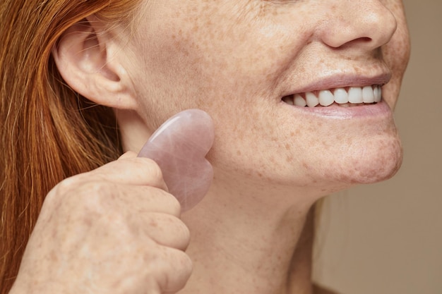 Close up of beautiful red haired woman with freckles smiling and doing face massage using roller cop...