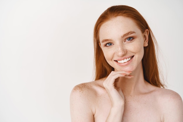 Photo close-up of beautiful redhead woman with pale skin, standing nude on white wall, smiling with perfect teeth, touching clean no makeup face