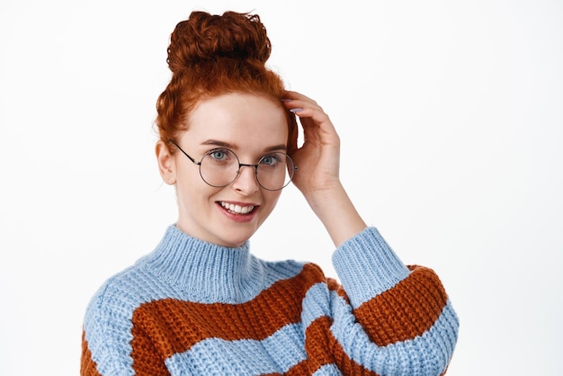 Close up of beautiful and romantic woman with ginger hair combed in hairbun wearing glasses touching hair and smiling flirty blushing cute standing against white background