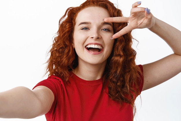Close up of beautiful smiling redhead woman showing v-sign and laughing, taking selfie, holding smartphone in stretched out hand, white wall