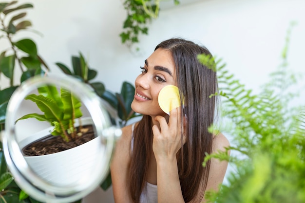 周りの植物と小さな鏡の前で自分自身の顔のマッサージをしている美しい女性をクローズアップ