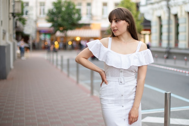 Primo piano di una bella donna con i capelli castani in un abito bianco che guarda di lato su uno sfondo di strada estiva. posto per il tuo design