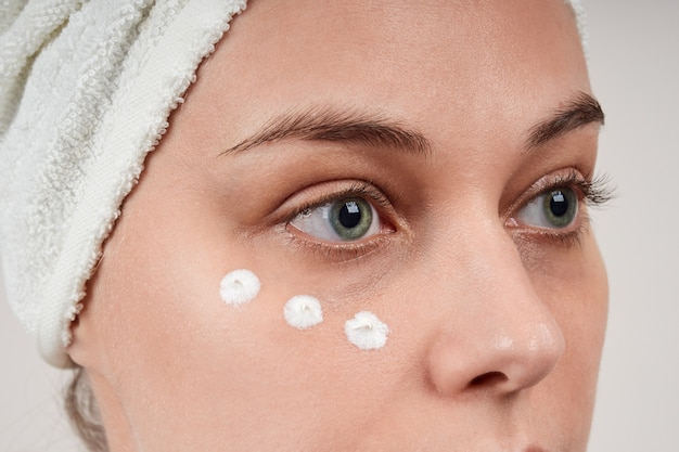 Primo piano di una bella giovane donna applica la crema sotto gli occhi, la cura della pelle del viso antirughe