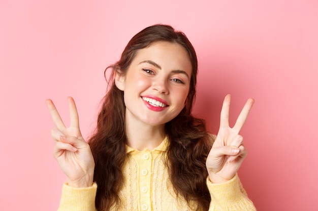 Close up of beautiful young woman showing peace v-sign and smiling happy at camera, wearing bright glam make up, standing against pink wall.