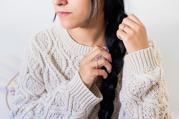 Close up of a beautiful young woman smiling and doing a braid in her hair in winter outfit at home