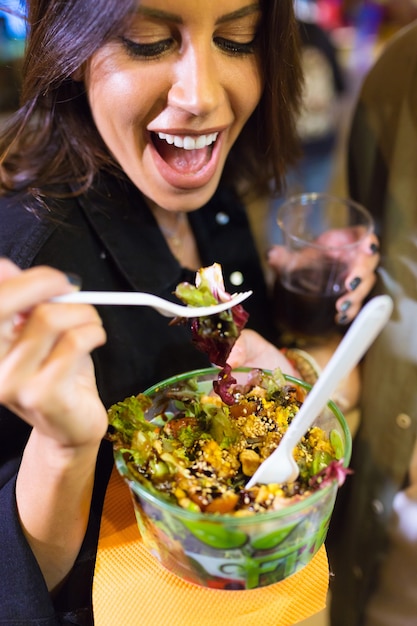 Close-up of beautiful young woman visiting eat market and eating colorful salad in the street.