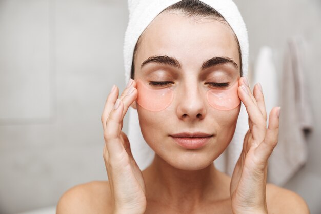 Photo close up of a beautiful young woman with towel on her head standing at the bathroom, applying eye patches