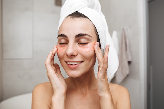 Close up of a beautiful young woman with towel on her head standing at the bathroom, applying eye patches