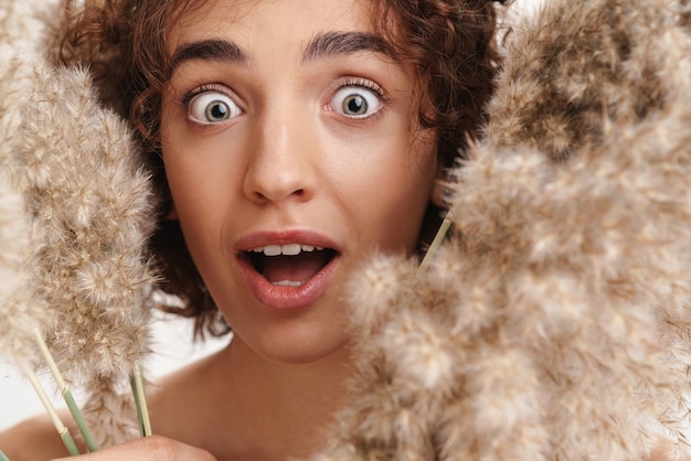 Close up beauty portrait of an attractive excited young topless woman with short curly brown hair isolated over white wall, holding dried herb