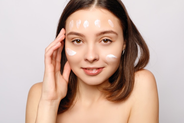 Close up beauty portrait of a laughing beautiful half naked woman applying face cream and looking away isolated over white background