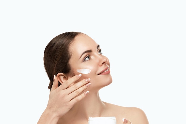 Close up beauty portrait of a laughing beautiful half naked woman applying face cream and looking away isolated over white background