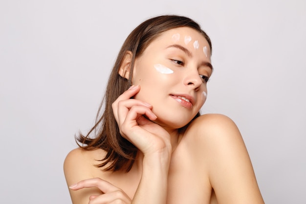 Close up beauty portrait of a laughing beautiful half naked woman applying face cream and looking away isolated over white wall