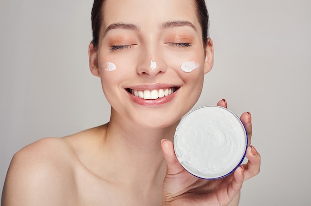 Photo close up beauty portrait of a laughing beautiful half naked woman with eyes closed holds in hand face cream.