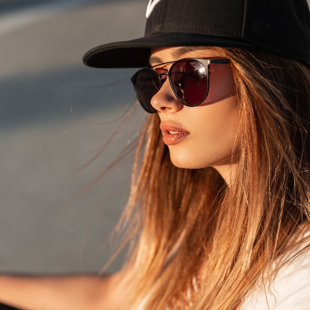 검은색 패션 선글라스와 야구 모자를 쓴 건강한 피부를 가진 섹시한 입술을 가진 꽤 매력적인 여성의 클로즈업 뷰티 초상화. 멋진 아름다운 소녀는 여름 일몰을 즐깁니다.