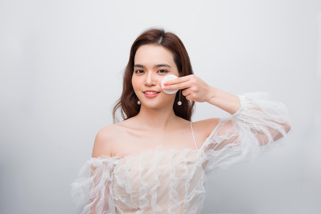 Close up beauty portrait of a smiling attractive woman cleaning her face with a cotton pad isolated over white background