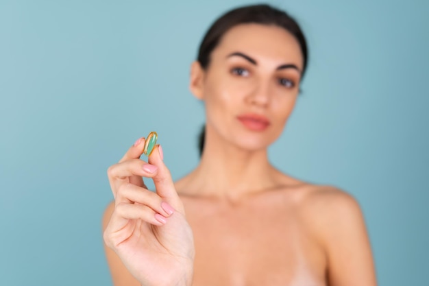 Close-up beauty portrait of a topless woman with perfect skin and natural make-up, holding a supplement, vitamin omega 3, fish oil, on a turquoise background