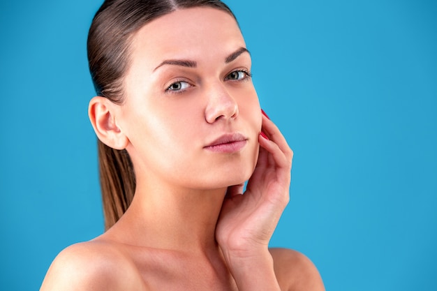 Close up Beauty portrait of young woman brunette smiling and touching her face on Blue wall. Perfect Fresh Skin. Youth and Skin Care Concept.