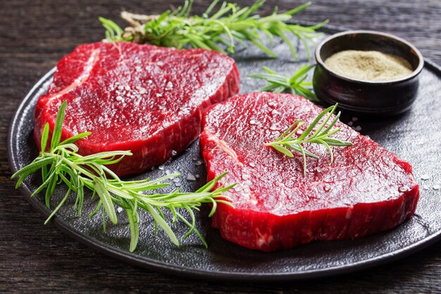 Close-up of beef steaks with rosemary and finely ground black peppercorn and salt on a black stone plate, landscape view from above