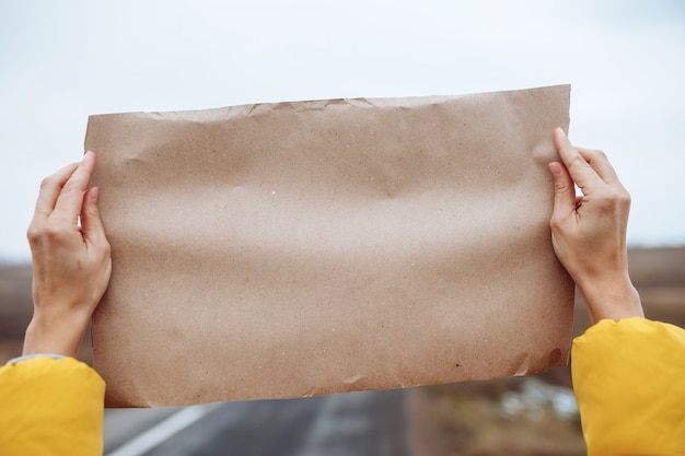 Close-up beeld van de handen van een vrouw die een gele jas draagt met een blanco posterpapier aan de kant van een lege winterweg.