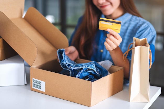 Close-up beeld van een jonge vrouw die creditcard vasthoudt en gebruikt voor online winkelen met boodschappentas en postpakketdoos met kleding op tafel